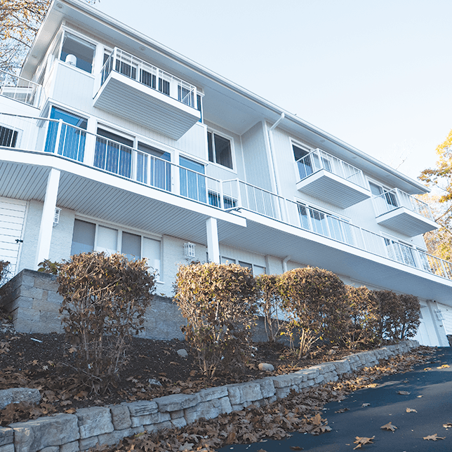 Multi-level white house with balconies, a stone retaining wall, and a sloped driveway lined with shrubs.